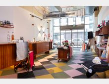 Ein heller, moderner Laden mit großen Glasfenstern, farbigem Schachbrettboden und Holzmöbeln. Das Interieur zeigt Regale mit Produkten, dekorative Accessoires und Tische mit Flaschen, Textilien und Dekorationsgegenständen.