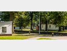 Eine grüne Parklandschaft mit hohen Bäumen umgibt ein kleines weißes Gebäude mit der Aufschrift „Schleuse Fürstenwalde/Spree“. Im Vordergrund ist ein schmaler Wasserkanal mit Pollern sichtbar, daneben ein gepflasterter Weg.