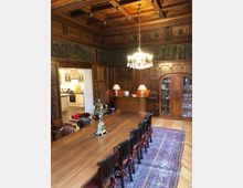 Holzgetäfelter Speisesaal mit einer langen Tafel, umgeben von Stühlen mit roten Polstern. Auf dem Tisch stehen Kerzenhalter, darüber hängt ein Kronleuchter. Im Hintergrund sind Glasvitrinen, ein gewebter Teppich und eine geöffnete Tür, die in eine helle Küche führt.