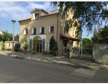 Gelb verputztes Wohn- oder Geschäftsgebäude mit roten Dachziegeln und mehreren Fenstern, eine zentrale Tür mit Rundbogen im Erdgeschoss. Der Eingang ist von Pflanzen in Töpfen umgeben, und rechts führt ein weißes Gittertor zu einem begrünten Innenhof.