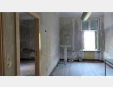 Ein leerer, verfallener Raum mit abblätterndem Putz an den Wänden, einem offenen Fenster mit einfachem Rahmen und einem kleinen weißen Stehtisch. Der Holzfußboden ist abgenutzt, und im angrenzenden Raum ist ein beschädigtes Mauerstück sichtbar.