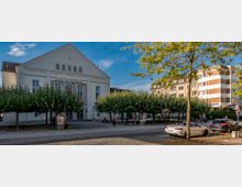 Außenansicht eines Kultur- und Festspielhauses mit heller Fassade, großen Fenstern und einem Giebeldach, umgeben von kleinen Bäumen in einem gepflasterten Platz. Im Vordergrund stehen geparkte Autos, und rechts sind moderne Wohngebäude zu sehen.