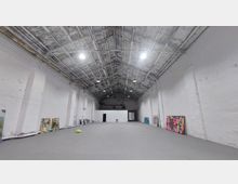 Ein großer, offener Raum mit hoher, sichtbarer Dachkonstruktion aus Metall und weißen Wänden, die teilweise durch vertikale Stützbalken unterteilt sind. Entlang der Wände sind bemalte Leinwände und Kunstwerke aufgestellt, der Boden ist grau und leer bis auf vereinzelte Gegenstände.