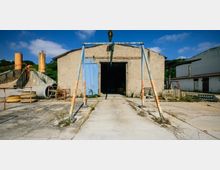 Industriegelände mit einer alten, beige-farbenen Lagerhalle, offenstehendem blauen Tor und rostigem Metallgerüst im Vordergrund. Im Hintergrund sind Betonröhren, gelbe Silos und weitere Gebäude zu sehen.