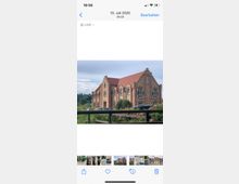 Backsteinbau im Stil der Renaissance mit markanten Treppengiebeln und roten Ziegeldächern, umgeben von blühenden Pflanzen und einer Rasenfläche; im Hintergrund eine leicht bewaldete Landschaft unter bewölktem Himmel.