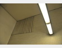 Decke und Wand eines modernen Innenraums mit weißen Ziegelwänden, vertikalen Lüftungsschlitzen und einer rechteckigen, leuchtenden Deckenleuchte.