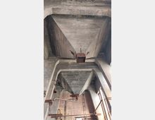 Industriegebäudeinnenraum mit massiver Betondecke und sichtbaren Trichterstrukturen. Im Hintergrund finden sich Ziegelwände, Fenster und einige rostige Rohre und metallische Strukturen.