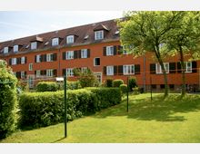 Mehrfamilienhaus mit orangefarbener Fassade und dunkelbraunen Fensterläden, umgeben von einer gepflegten Grünanlage mit Hecken, Bäumen und einem Rasen. Im Vordergrund befinden sich Wäscheleinen und Stangen.