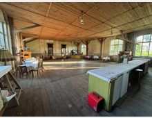 Großer, rustikaler Saal mit Holzfußboden und Holzdecke, sichtbaren Stützbalken und mehreren hängenden Lampen. Entlang der Wände stehen kleine Tische und Stühle, im Vordergrund eine lange Theke; große Sprossenfenster lassen viel Tageslicht herein und geben den Blick ins Grüne frei.