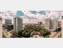 Panoramablick auf ein städtisches Viertel mit mehreren hohen Wohn- und Bürohochhäusern, einem langgestreckten Gebäuderiegel und breiten Straßen. Im Vordergrund liegt ein dicht bewachsener Park mit einem halbkreisförmigen, monumentalen Säulenbau und einem Platz.