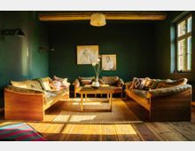 Wohnzimmer mit dunkelgrünen Wänden, Holzbalkendecke und einem großen Fenster, das Tageslicht hereinlässt. In der Mitte stehen ein Holztisch und ein Teppich, umgeben von drei gemütlichen Sofas aus Holz mit beigen Polstern und bunten Kissen; an der Wand hängen zwei Zeichnungen.