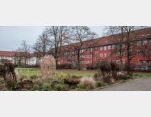 Rote Backsteingebäude mit weißen Fenstern und roten Ziegeldächern, im Vordergrund eine Grünfläche mit kahlen Bäumen und winterlicher Vegetation. Der Himmel ist bewölkt und der Weg im rechten Bildbereich führt durch die Grünanlage.