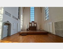 Heller Kirchen- oder Kapelleninnenraum mit hoher Gewölbedecke, großem hohen Fenster und einer Orgel auf einer erhöhten Holzplattform. Weiße Wände mit Akustikpaneelen und Holzboden prägen den Raum, seitlich sind weitere hohe Fenster und eine Tür sichtbar.