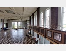 Großer, heller Saal mit hoher Decke, bodentiefen Fenstern und einer Galerie mit Holzgittergeländer an einer Backsteinwand. Der Boden ist mit geometrischen Mustern aus Holzparkett gestaltet, und die Einrichtung wirkt minimalistisch.