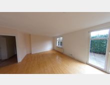 Ein leerer Wohnraum mit hellen Wänden und einem Laminatboden aus Holz. Der Raum verfügt über ein großes Fenster, eine Terrassentür mit Blick ins Grüne und einen Heizkörper unter dem Fenster; eine Tür führt in einen angrenzenden Bereich.