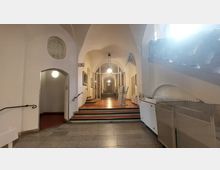 Innenansicht eines historischen Gebäudes mit einem Flur aus weiß verputzten Wänden und gewölbten Decken. Im Zentrum befindet sich eine Treppe mit hölzernen Stufen, flankiert von Handläufen, und elektrische Infopulte stehen entlang der Wände.