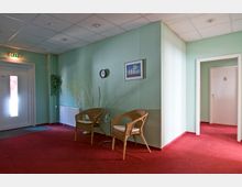 Flur mit mintgrünen Wänden, rotem Teppichboden und einer Sitzgruppe aus zwei geflochtenen Stühlen und einem kleinen Glastisch. Links eine Tür mit einem leuchtenden Notausgangsschild darüber, rechts eine weiße Tür mit der Nummer 5.