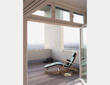 Offene Terrasse mit Holzboden und einer verstellbaren Liege mit grünen Polstern, Blick auf eine städtische Landschaft im Hintergrund. Glasfenster und -türen rahmen den Zugang zur Terrasse ein.