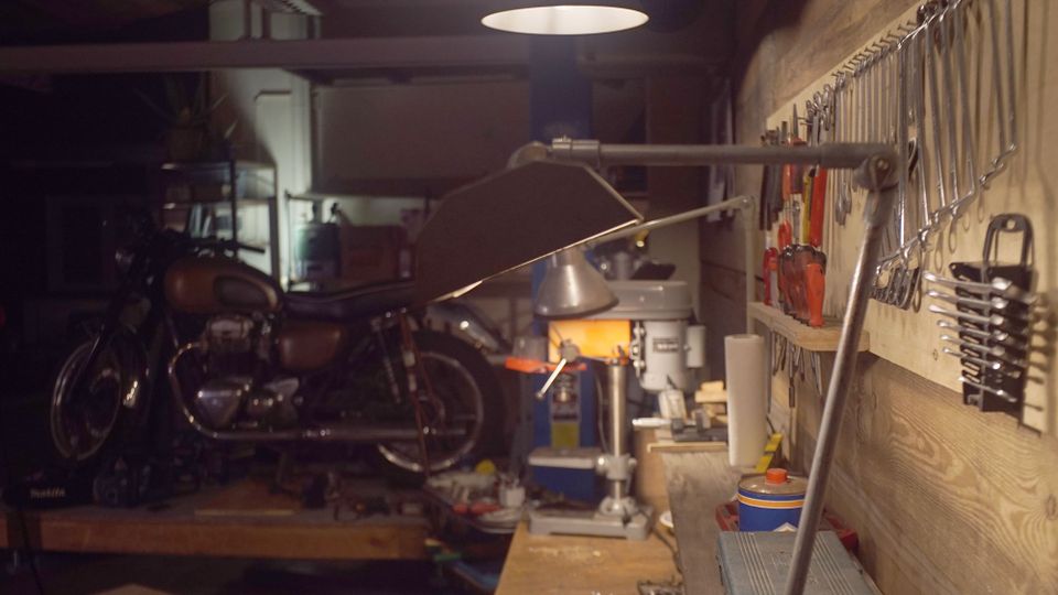 Innenansicht einer Werkstatt oder Garage mit Werkbank im Vordergrund und einer Werkzeugwand mit aufgehängten Schraubenschlüsseln und Zangen. Im Hintergrund stehen ein Motorrad und eine Ständerbohrmaschine unter gedämpfter Deckenbeleuchtung.
