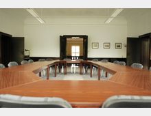 Konferenzraum mit U-förmiger Anordnung aus hölzernen Tischen und gepolsterten Stühlen. Im Hintergrund befindet sich eine offene doppelflügelige Tür, die in einen hell beleuchteten Raum führt, und an den Wänden hängen gerahmte Bilder.