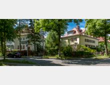 Straßenszene in einem Wohngebiet mit zwei großen, hell gestrichenen Häusern im Stil der klassischen Architektur, umgeben von Bäumen und üppigem Grün. Die gepflasterte Straße verläuft am Vordergrund, und ein Straßenschild zeigt "Sophie-Charlotten-Straße".