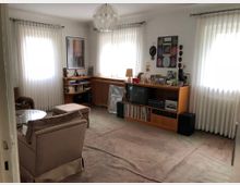 Wohnzimmer mit beigefarbenem Teppichboden, zwei hellbraunen Sesseln, einem Glastisch sowie einer Holzanrichte, die Bücher, eine Stereoanlage, Schallplatten und Fotos zeigt. Drei Fenster mit weißen Gardinen lassen Tageslicht herein, und an den Wänden hängen Bilder und Dekorationen.