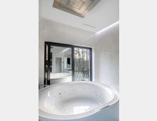 Luxuriöses Badezimmer mit freistehender Whirlpool-Badewanne, großer Glas-Schiebetür, die auf einen Balkon mit dekorativem Geländer führt, sowie einer beleuchteten Deckeninstallation aus Edelstahl. Im Hintergrund sind Spiegel und Fenster mit Blick auf Bäume sichtbar.