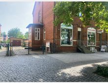 Backstein-Gebäude mit Geschäften im Erdgeschoss, darunter ein Fotogeschäft und ein Reisebüro. Davor ein gepflasterter Vorplatz mit einem Metalltor, Bäumen im Vordergrund und einer Garage im Hintergrund.