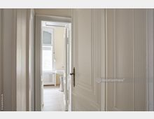 Blick durch eine geöffnete weiße Tür in ein helles Badezimmer mit beigefarbenen Wänden, Fliesenboden, einem Waschbecken und einem großen Fenster mit halb geschlossenen Rollläden.