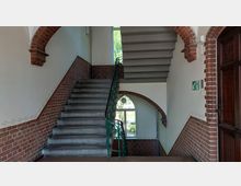 Treppenhaus in einem historischen Gebäude mit gemauerten Wänden und Ziegelbögen, Metallgeländer und Blick durch ein halbkreisförmiges Fenster nach draußen. Die Treppe teilt sich auf zwei Ebenen, und ein Notausgangsschild ist an der Wand angebracht.