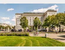 Großes, helles Stein-Gebäude im Stil der frühen Moderne mit vielen Fenstern und einem hohen Durchgang im Erdgeschoss, davor eine breite Straße mit Haltestellen und Bäumen. Im Vordergrund liegt eine Rasenfläche mit Blumenbeet und Radweg, umgeben von städtischer Bebauung.