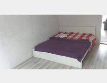 Ein schlichtes Schlafzimmer mit einem weißen Bettgestell, einer rot-weiß karierten Bettdecke und einer lila Decke darüber. Der Raum hat helle Wände und einen hellen Laminatboden; rechts im Bild ist eine offene Tür zu erkennen.