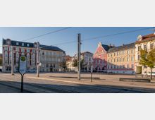 Platz mit historischen Wohn- und Geschäftshäusern im Hintergrund, davon ein auffälliges rosa Gebäude mit Ziergiebel; im Vordergrund Straßenbahnschienen, eine Haltestellenschild mit der Aufschrift "Dresdener Platz" und vereinzelte Bäume.