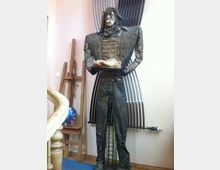 Eine Skulptur eines stehenden Mannes mit detailreichen Gesichtszügen und übergroßen, eckigen Schultern. Die Figur hält ein Tablett mit einem Brotstück, steht vor einer Wand mit gebogenen Metallstangen und ist in einem künstlerischen Raum platziert, erkennbar an Staffeleien und einer Malpalette im Hintergrund.