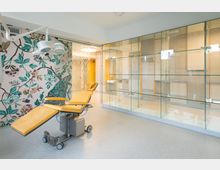 Behandlungsraum mit einer gelben medizinischen Liege, einer Deckenleuchte und Wandverkleidungen mit floralem Mosaikmuster. Auf der rechten Seite befinden sich gläserne Vitrinen vor einer hellen Wand.