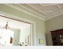 Innenansicht eines Wohnzimmers mit Stuckdecke und pastellgrünen Wänden. Sichtbar sind ein Holzelement der Tür, eine Hängelampe, ein Holzschrank mit Glastüren sowie Dekorationen und Möbelstücke an den Wänden und vor einem großen Fenster.