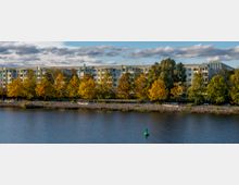 Wohngebäude entlang eines Ufers mit herbstlich gefärbten Bäumen im Vordergrund. Der Fluss im Vordergrund wird von einer Steinböschung gesäumt, und im Wasser ist eine grüne Boje zu sehen.