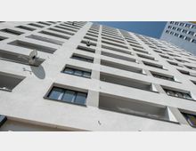 Wohnhochhaus mit weißer Fassade, mehreren Fensterelementen und Balkonen, aufgenommen aus der Froschperspektive. An der Fassade sind Satellitenschüsseln angebracht, und der Himmel ist klar und blau.