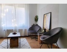 Helles Wohnzimmer mit zwei grauen, runden Sesseln und einem rechteckigen Couchtisch mit einem geflochtenen Korb darunter. Im Hintergrund ein großes Fenster mit weißen Vorhängen und eine Wand mit einem gerahmten Bild und Dekorationen.