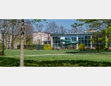 Außenansicht einer Schwimmhalle mit einer großen Glasfassade und einer grünen Röhrenrutsche an der Seite, umgeben von einer Grünfläche mit Bäumen und Wegen. Im Hintergrund sind Wohnhäuser und blauer Himmel sichtbar.