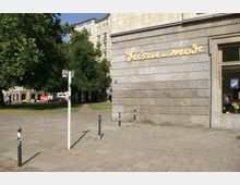 Außenansicht eines Gebäudes mit beiger Steinwand und der Aufschrift „Frisur u. Mode“ in gelber Schrift. Im Vordergrund ist ein gepflasterter Platz mit Pollern und einem Schild, dahinter ein kleiner Park mit Bäumen und mehrstöckigen Wohnhäusern im Hintergrund.