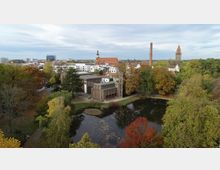 Luftaufnahme eines städtischen Parks mit einem Teich im Vordergrund, umgeben von herbstlich gefärbten Bäumen. Im Hintergrund sind ein backsteinernes Gebäude mit modernem Anbau, ein hoher Schornstein und eine Kirche mit rotem Dach sowie ein Glockenturm zu sehen, eingebettet in eine Stadtlandschaft.