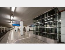 U-Bahn-Station mit einem gebogenen Bahnsteig, beleuchtet durch Reihen von Deckenleuchten. Im Vordergrund ist ein verglaster Aufzugsschacht sichtbar, und im Hintergrund sind weiße Säulen, Anzeigetafeln und eine Rolltreppe erkennbar.