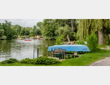 Seeufer mit einer blauen umgedrehten Bootsschale auf einer kleinen Wiese, daneben eine rustikale Bank. Im Hintergrund ist ein ruhiger Fluss mit Bäumen, Schilf und kleinen Gebäuden am Ufer sichtbar.
