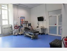 Ein heller Behandlungsraum mit blauen Bodenfliesen, einer Krankenliege in der Mitte, medizinischen Geräten und Monitoren an der Wand sowie Fenstern und einer Glastür im Hintergrund. Ein Feuerlöscher steht rechts im Vordergrund.