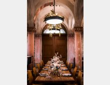 Eleganter Speisesaal mit einer langen, gedeckten Holztafel, dekoriert mit Blumenarrangements und Kerzen; umgeben von hohen, rosa gefärbten Säulen und Rundbögen in einem historischen Gebäude mit einer hölzernen Doppeltür im Hintergrund.