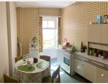 Küche im Retro-Stil mit gemusterten Tapeten in Gelb- und Brauntönen, einem alten Gasherd, weißen Schränken und einem runden Esstisch mit kariertem Tischtuch. Ein Fenster mit bunten Vorhängen lässt Tageslicht herein, und der Boden ist mit hellgrünem Linoleum bedeckt.