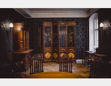 Ein eleganter Raum mit dunkler Tapete und antiken Möbeln, darunter ein reich verzierter hölzerner Schrank und ein Paravent mit asiatischen Motiven. Der Holzboden und die verzierten Stühle betonen die historische Atmosphäre.