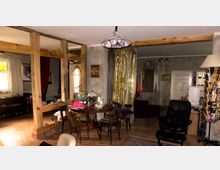 Wohnzimmer mit rustikalem Charme, geprägt von einer Holzbalkenkonstruktion, goldenen Vorhängen und wechselnden Stilelementen wie einem runden Holztisch mit Stühlen, einer Stehlampe und dekorativen Akzenten wie einem Globus und Kunstwerken. Der Raum hat teils freigelegte Wände und einen hellen Fliesenboden.