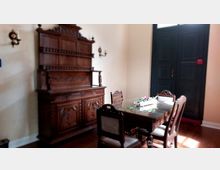 Ein Raum mit antiken Möbeln, darunter ein reich verzierter Buffet-Schrank aus dunklem Holz und ein Esstisch mit passenden Stühlen. Auf dem Tisch liegt eine dekorative Blumengesteck, und im Hintergrund befindet sich eine dunkle Holztür.